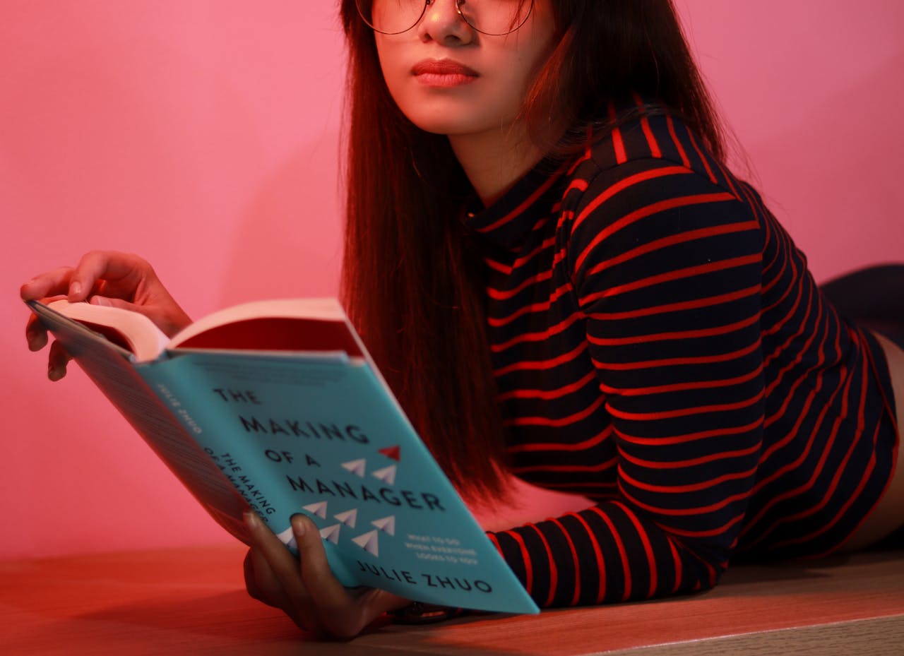 A woman lying on a pink background reading a book called The Making of a Manager.